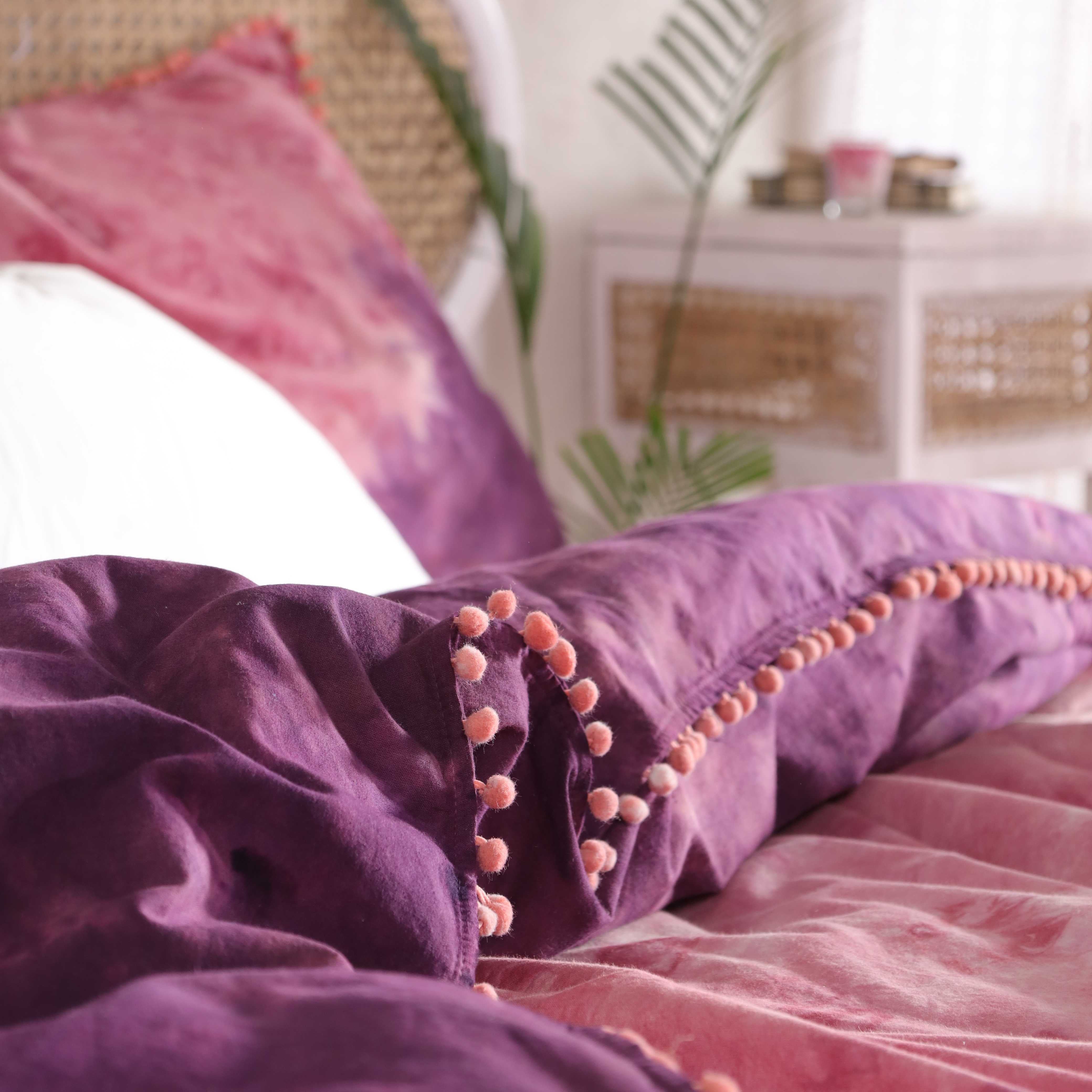 Two matching purple and pink tie dye pillow shams with pom pom borders resting on a bed.