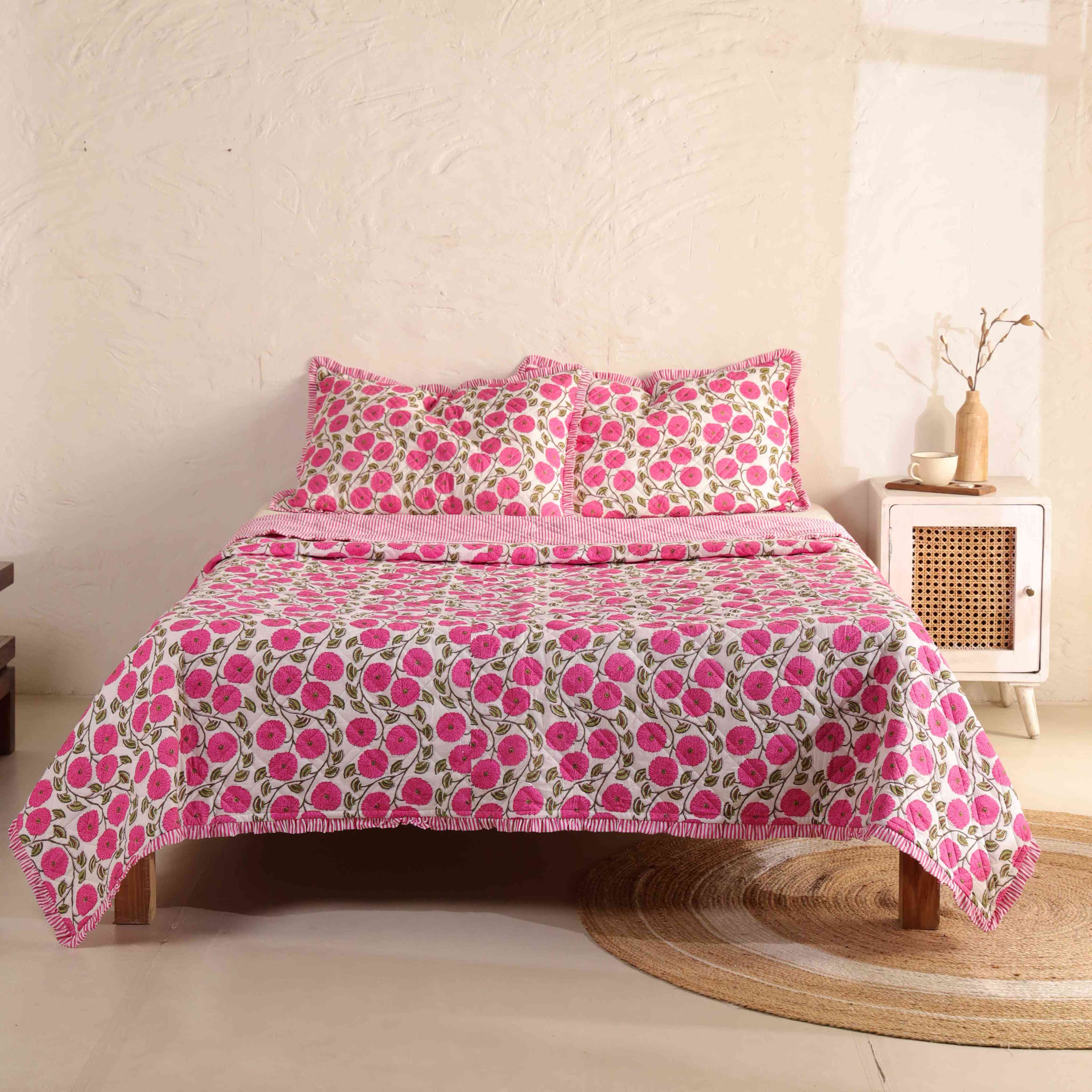 3-piece pink floral quilt set with ruffled edges on a wooden bed in a bright bedroom.