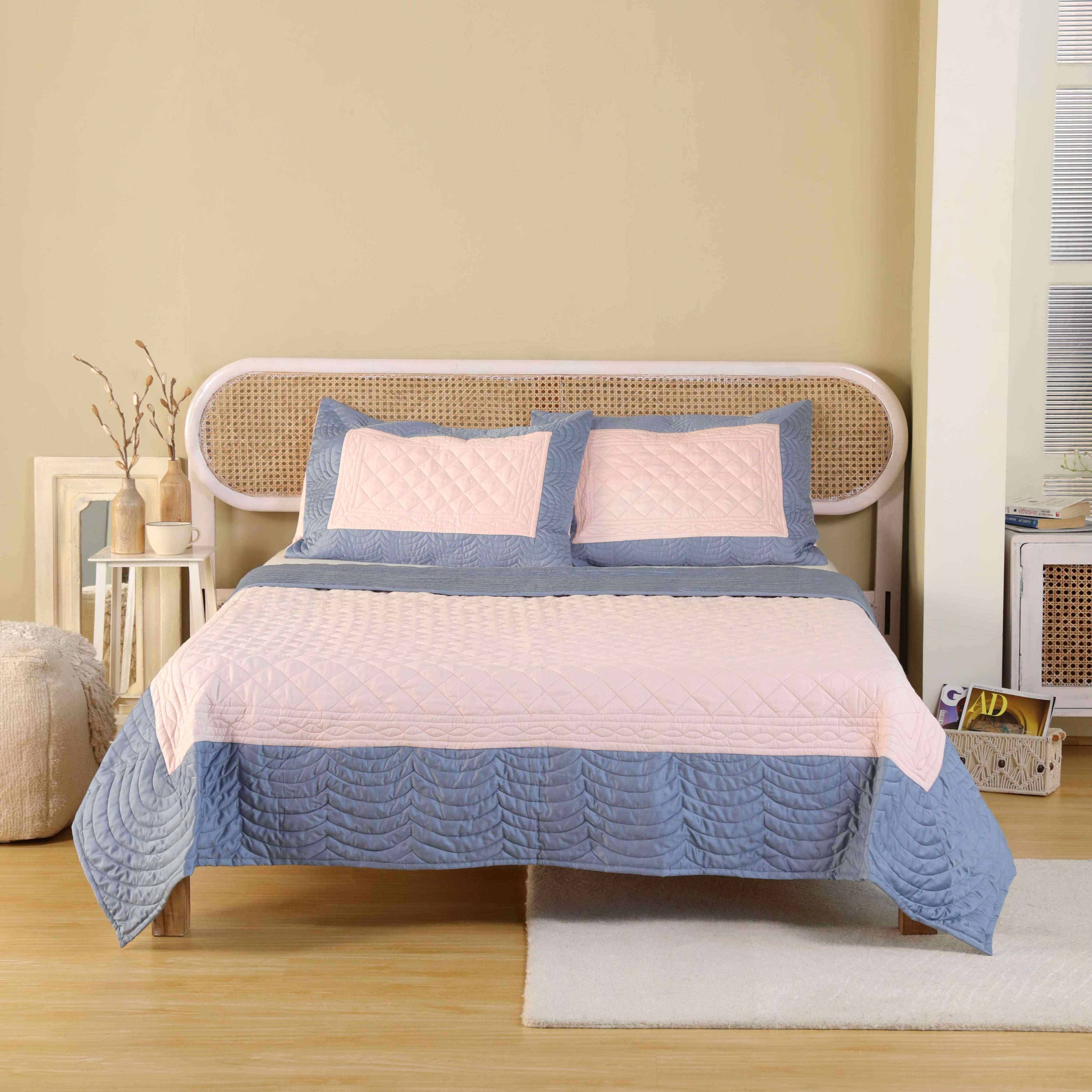 A dusty blue and blush pink color-block satin quilt set on a bed with a rattan headboard.