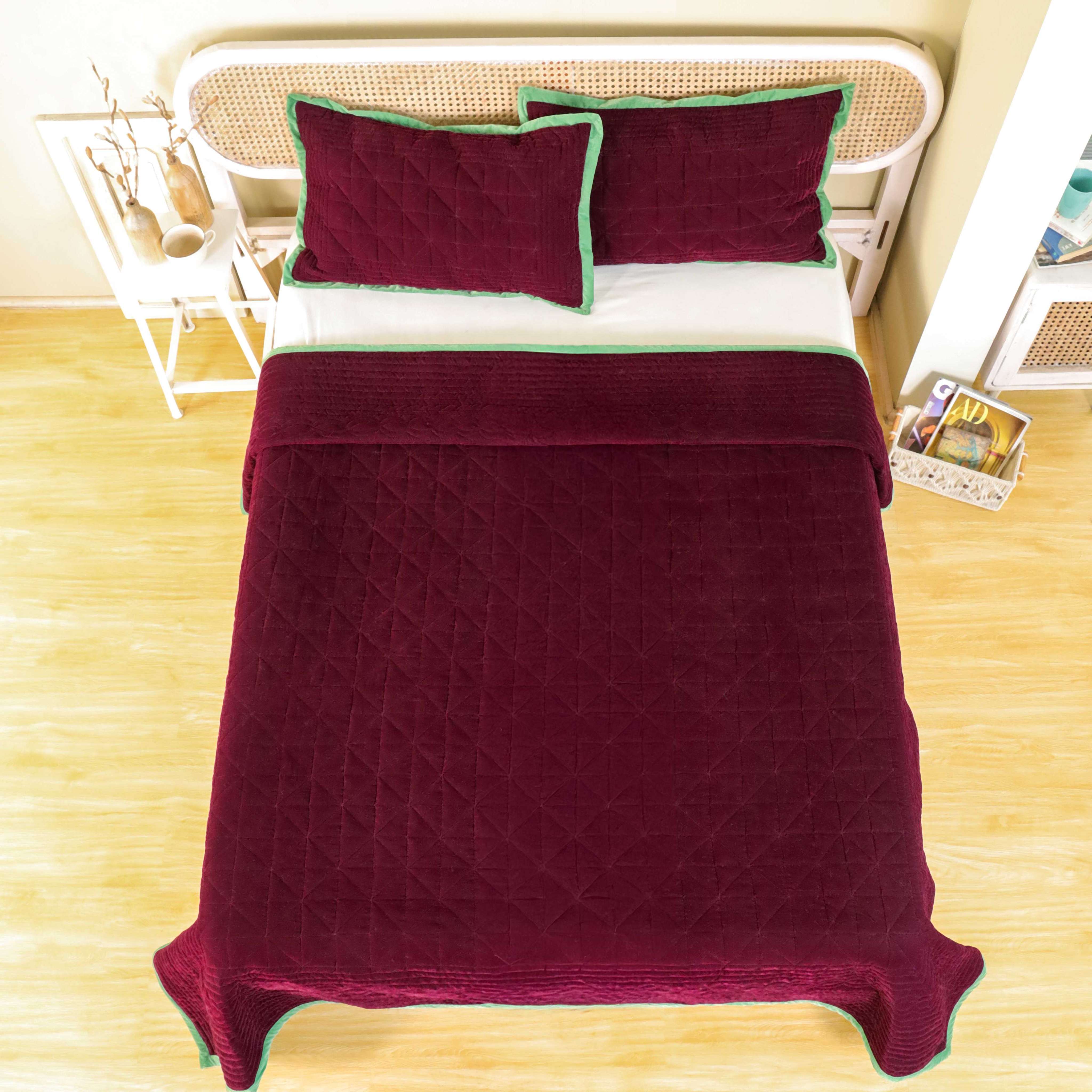 Maroon velvet duvet cover with green trim and diamond quilting on a bed with a cane headboard.